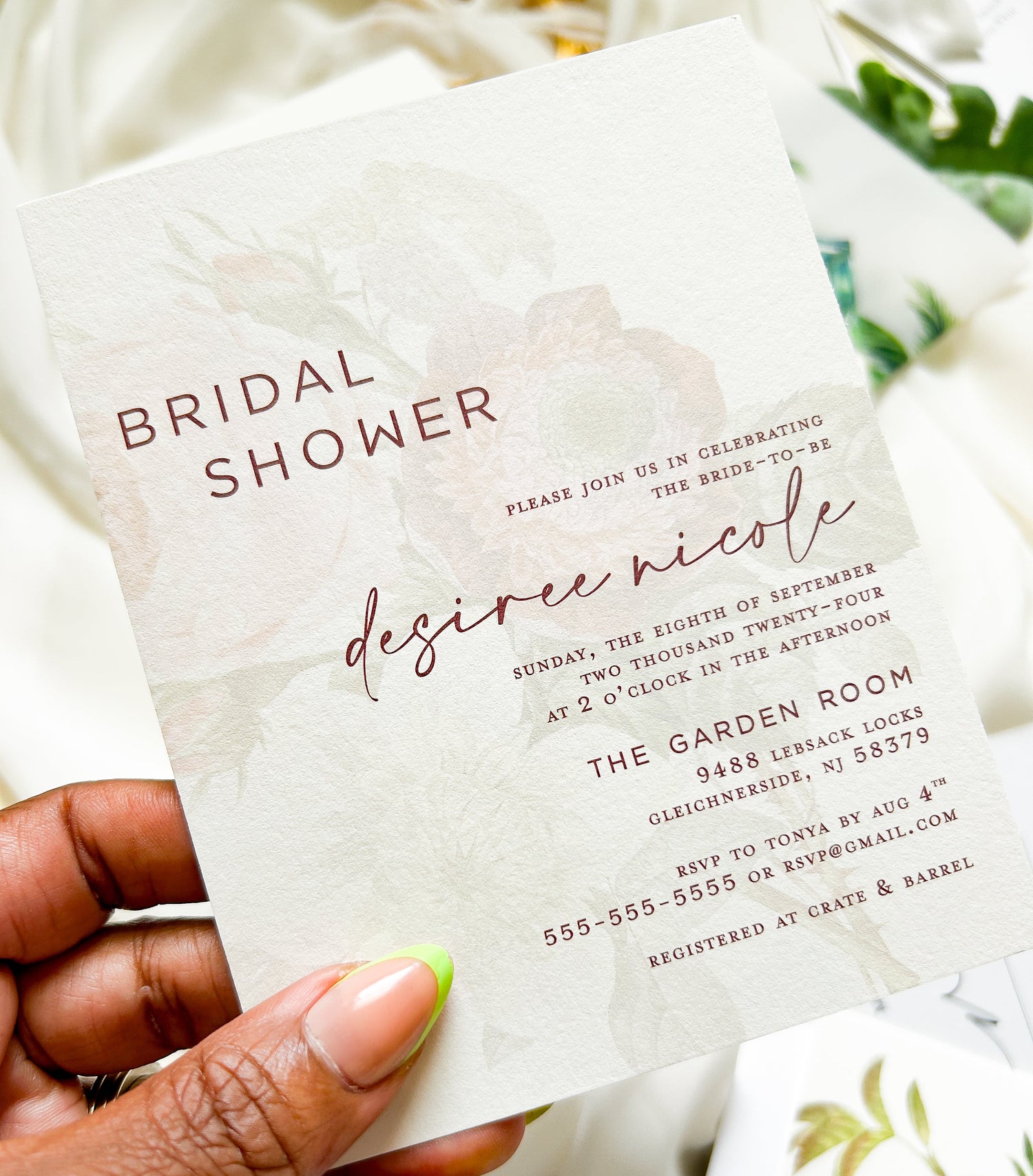 A hand holds a bridal shower invitation card with a soft blush pink floral design and rose-gold text. The card reads 'Bridal Shower' and 'Desiree Nicole' with event details below, set against a softly blurred background with white fabric and greenery.