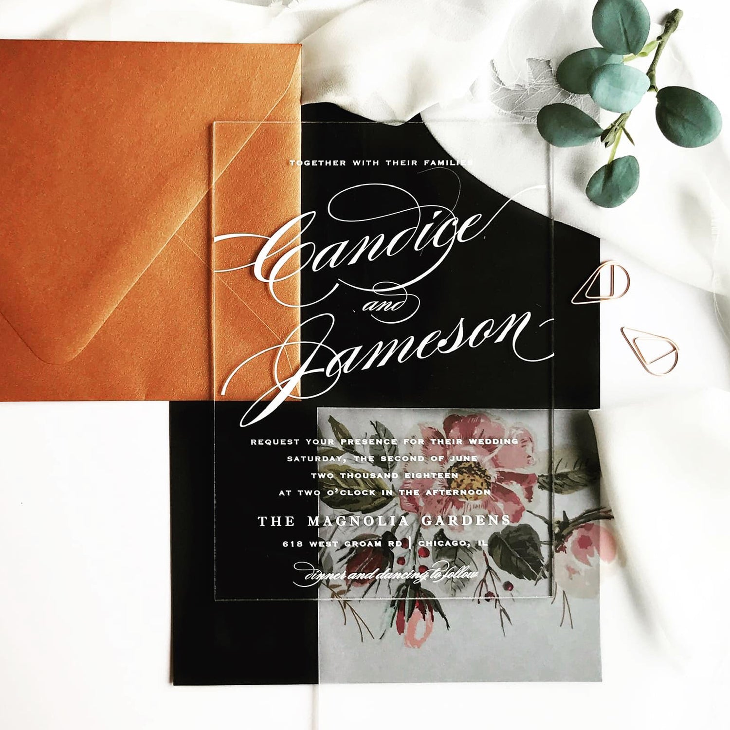 A styled flat-lay of a wedding invitation card made of clear acrylic with white script lettering, placed over a black and white floral design. An orange-brown envelope is visible on the left, with white fabric, green eucalyptus leaves, and small rose gold paperclips around it.