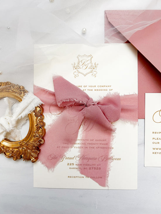 A wedding invitation suite featuring a cream card with gold foil detailing, a dusty pink watercolor bow, a matching pink envelope, and a small white card, styled on a light gray surface with a gold mirror and white pearls.