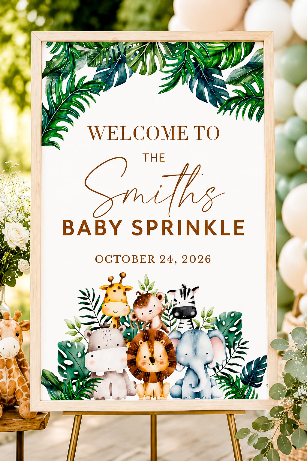 A framed baby shower sign on an easel with tropical leaf borders, the text 'Welcome to the Smiths Baby Sprinkle October 24, 2026,' and cartoon safari animals including a lion, elephant, zebra, giraffe, and hippo. The sign is photographed in a real event setting with balloons and floral decor in the background.