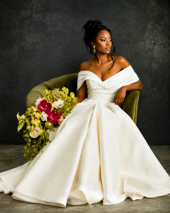 A woman wearing an off-the-shoulder white satin wedding gown with a full skirt, seated on a green velvet armchair against a dark textured wall, holding a large floral bouquet.